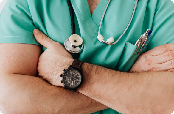 Um médico de braços cruzados, utilizando um relógio preto, camiseta verde e um estetoscópio em volta do pescoço 